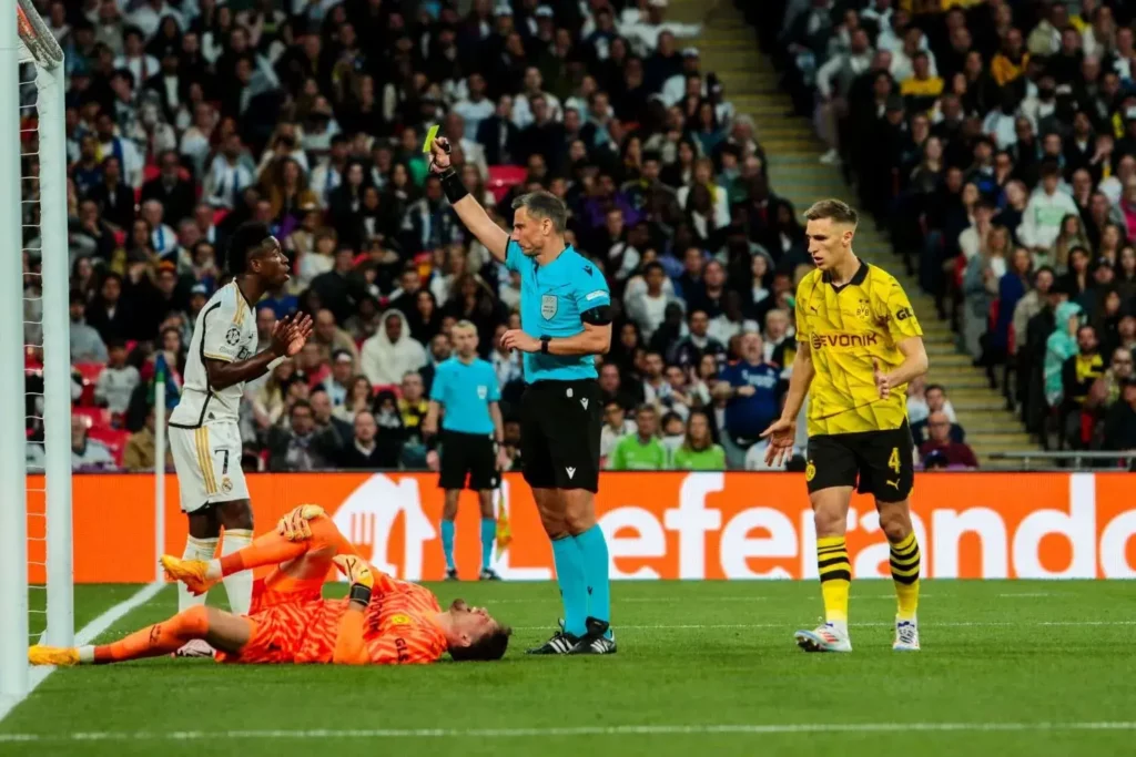Gregor Kobel von Dortmund liegt verletzt auf dem Boden und der Schiedsrichter zeigt Vinícius Junior von Real Madrid die gelbe Karte
