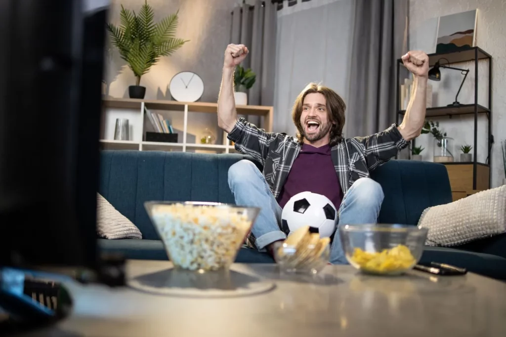 Jubelnder Fußballfan beim Fußball gucken vor dem Fernseher und mit einem Ball zwischen seinen Beinen