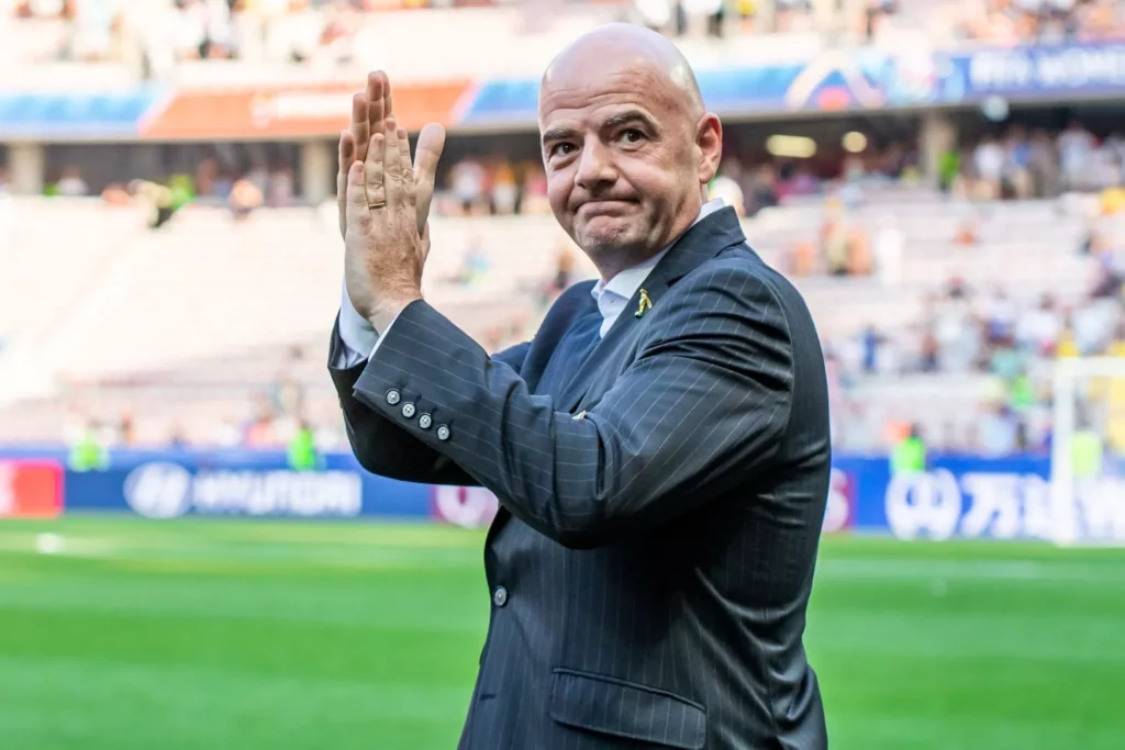 Gianni Infantino geht mit erhobenen Händen und leichtem Applaus über ein Fußballfeld